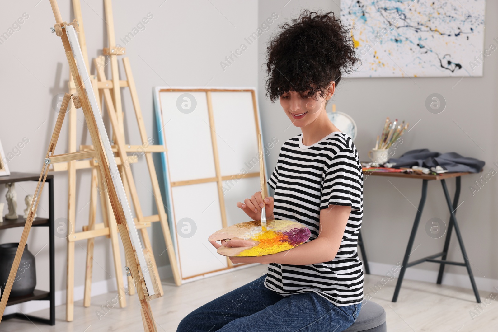 Photo of Young woman mixing paints on palette with brush near easel in studio