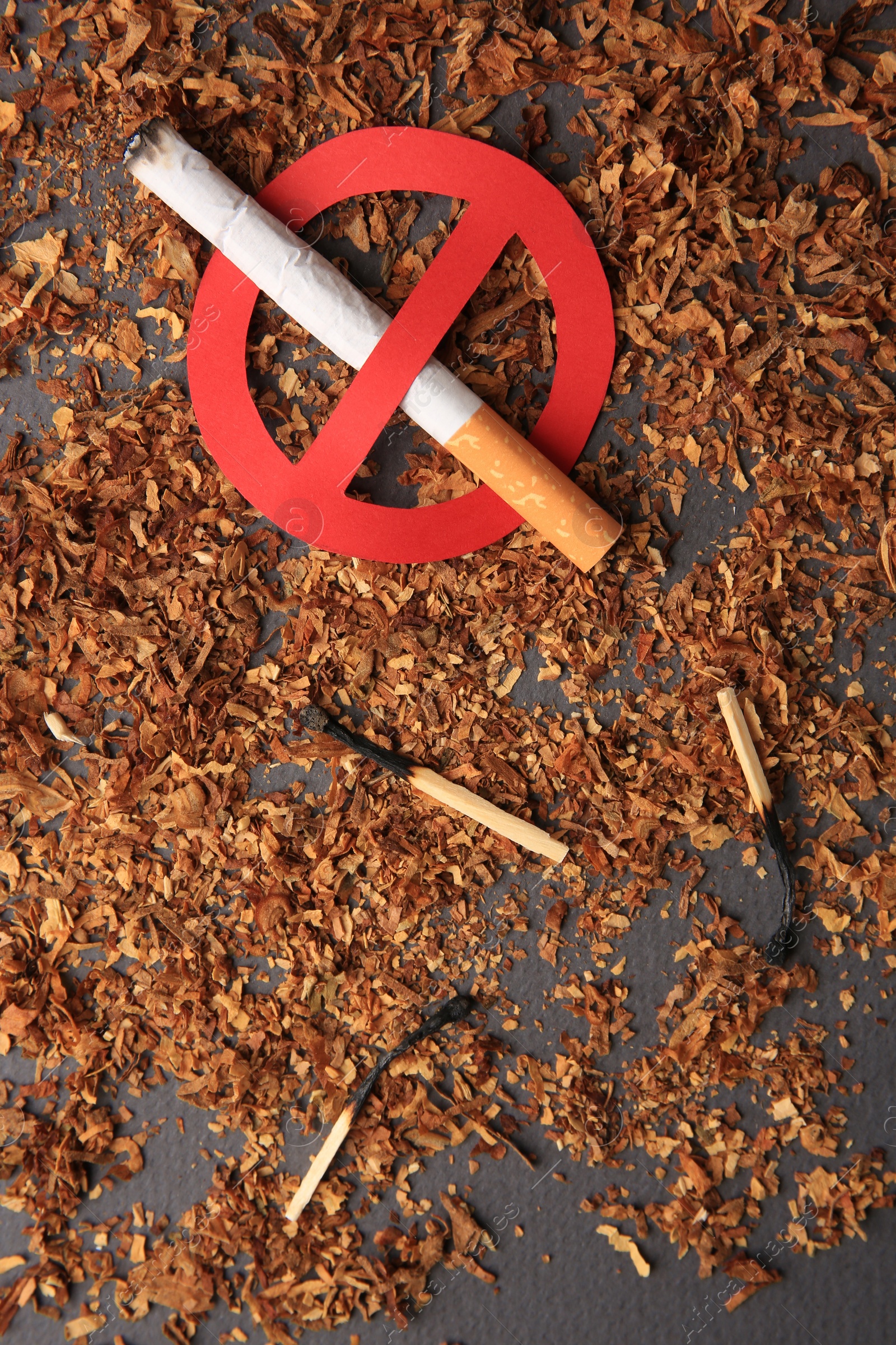 Photo of Cigarette with prohibition sign, burnt matches and dry tobacco on dark grey background, flat lay. Quitting smoking concept