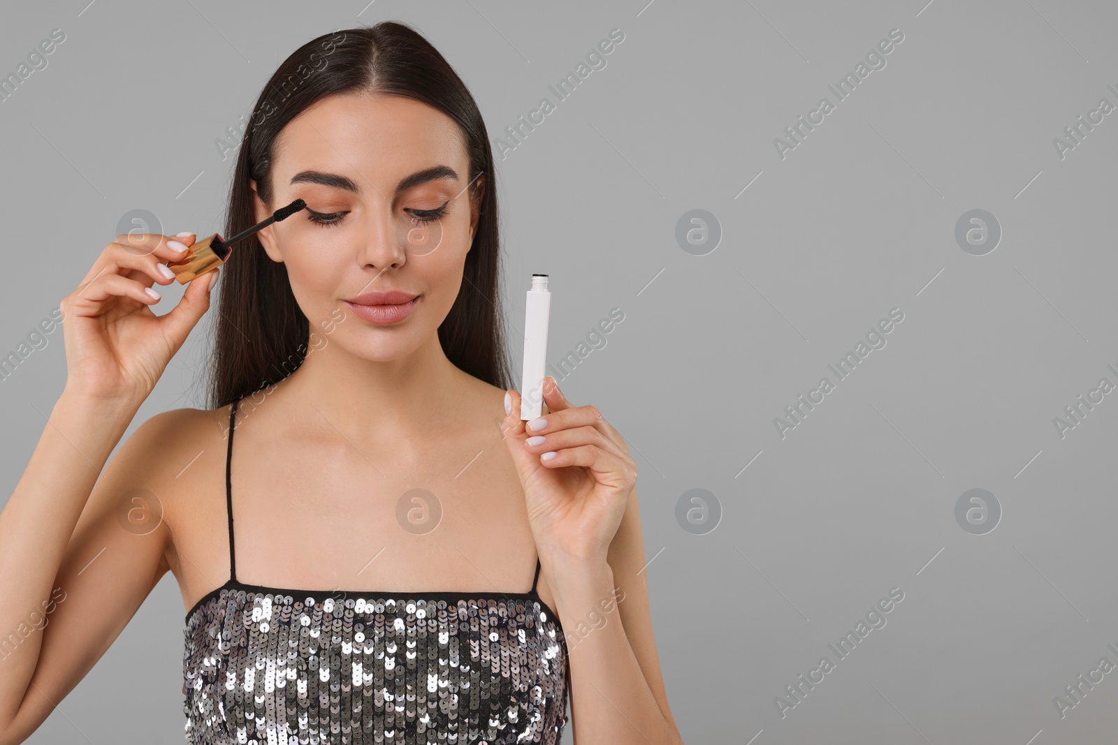 Photo of Beautiful young woman applying mascara on grey background, space for text