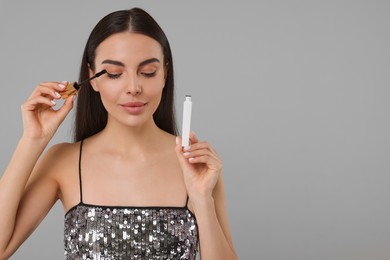 Photo of Beautiful young woman applying mascara on grey background, space for text