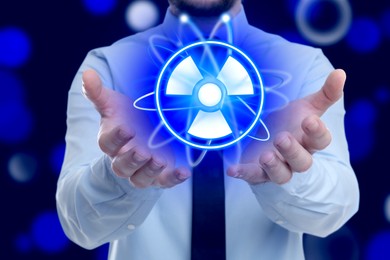 Man holding atom symbol with radiation warning sign on dark blue background, closeup. Bokeh effect