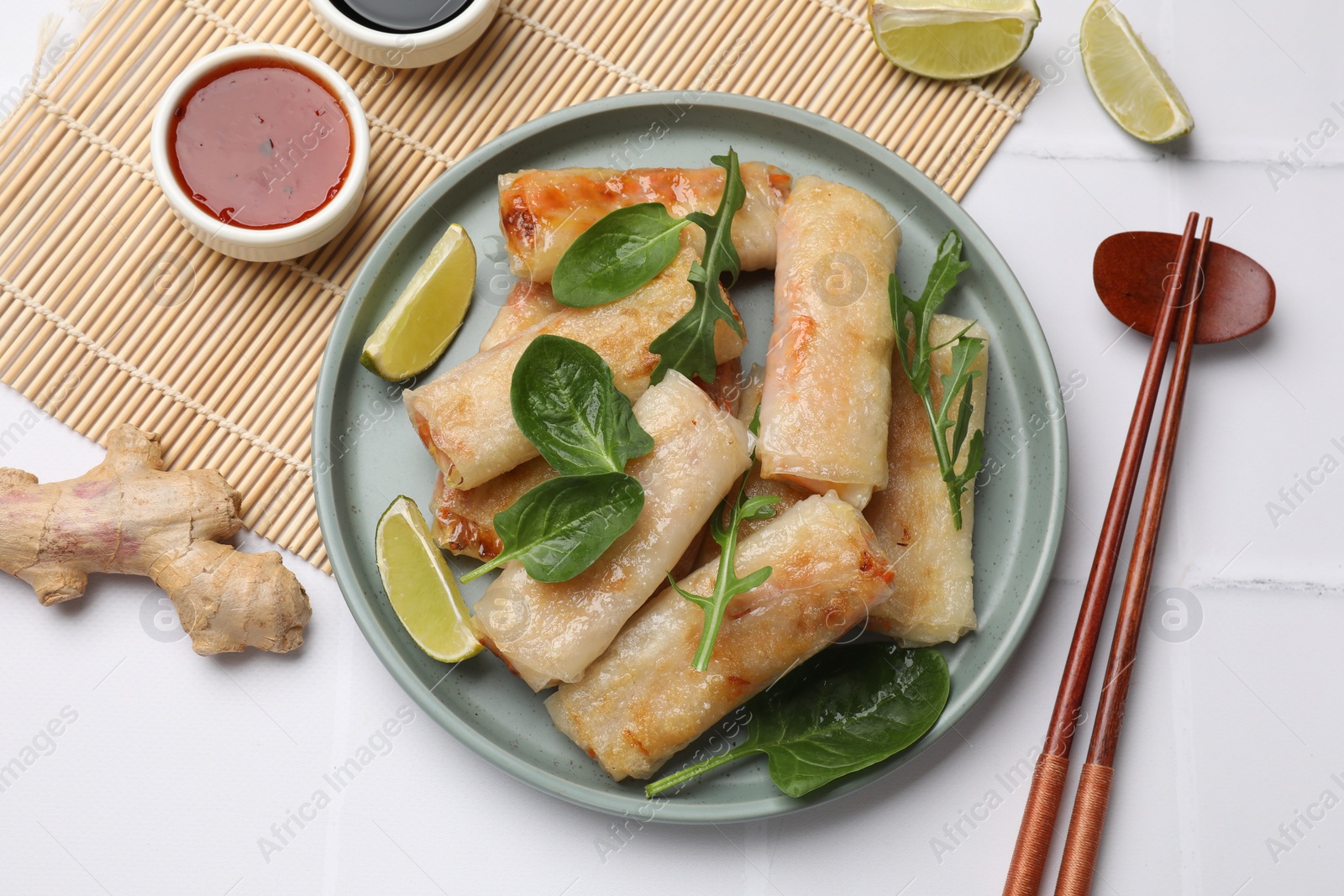 Photo of Plate with tasty fried spring rolls, spinach, arugula and lime served on white tiled table, flat lay