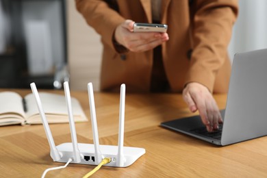 Photo of Woman with smartphone and laptop connecting to internet via Wi-Fi router at table indoors, selective focus