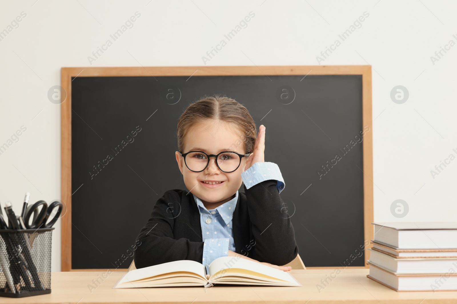 Photo of Happy little school child raising hand while sitting at desk with books near chalkboard