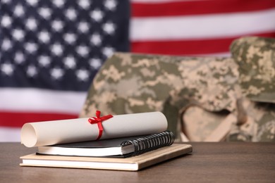 Notebooks and diploma on wooden table, chair with soldier uniform against flag of United States indoors. Military education