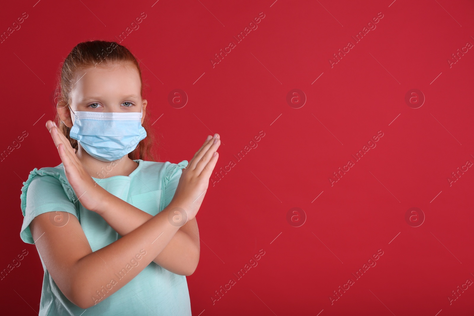Photo of Little girl in protective mask showing stop gesture on red background, space for text. Prevent spreading of coronavirus