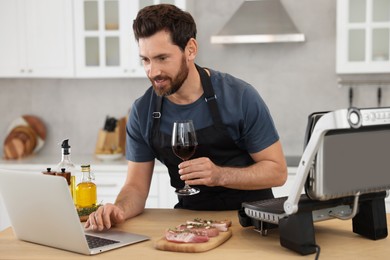 Photo of Man with glass of wine making dinner while watching online cooking course via laptop in kitchen