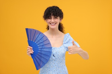 Photo of Happy woman holding hand fan and showing thumb up on orange background