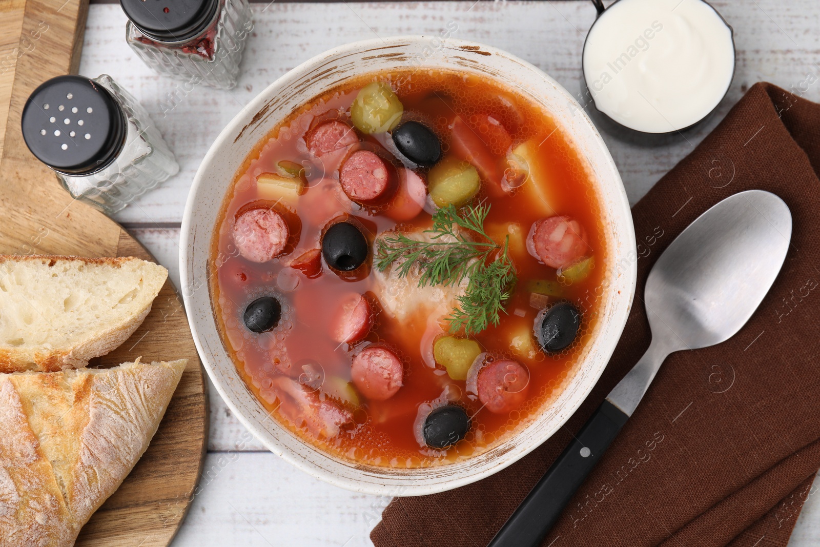 Photo of Meat solyanka soup with thin dry smoked sausages served on white wooden table, flat lay