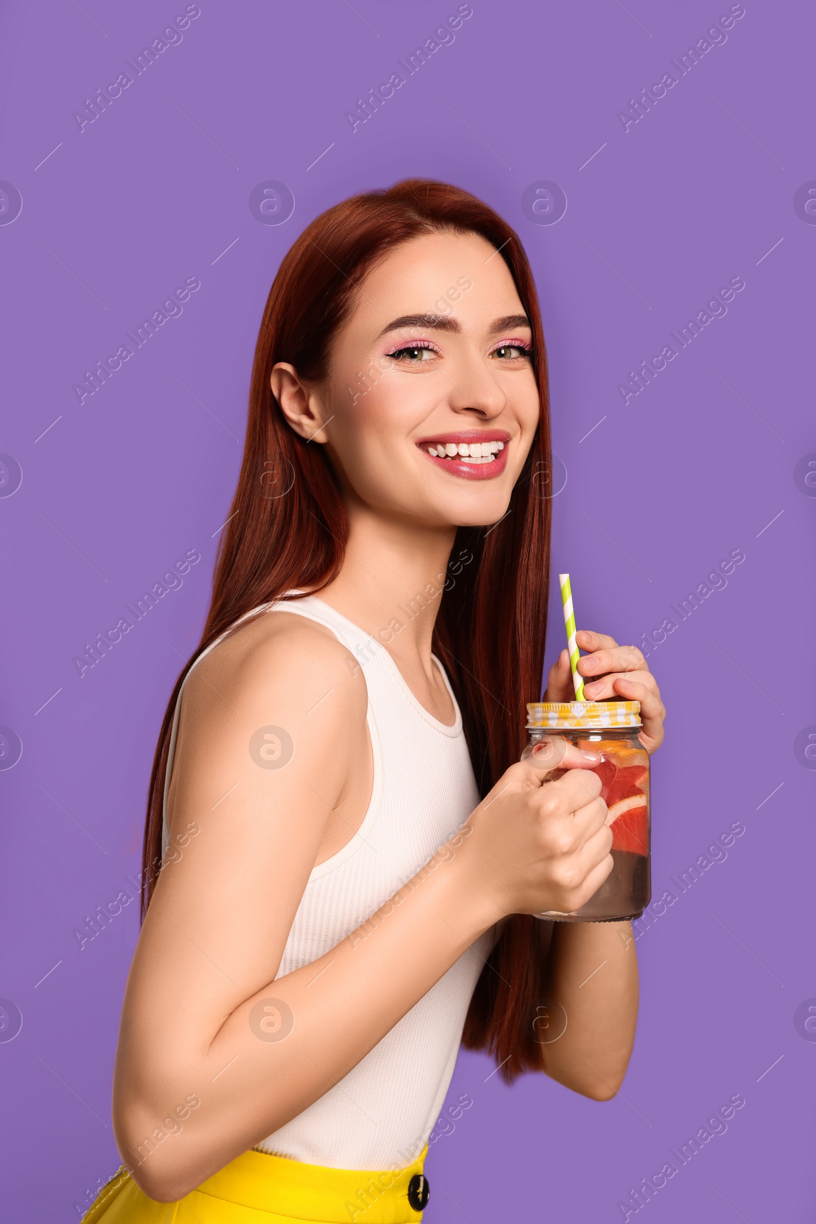 Photo of Happy woman with red dyed hair and refreshing grapefruit drink on purple background