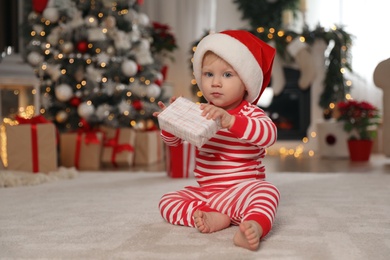 Photo of Cute baby in Christmas pajamas and Santa hat holding gift box at home