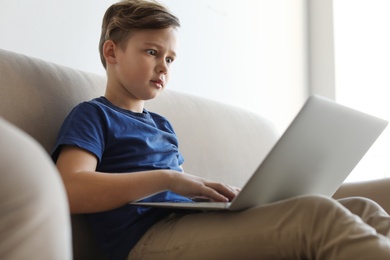 Photo of Shocked little child with laptop on sofa in room. Danger of internet