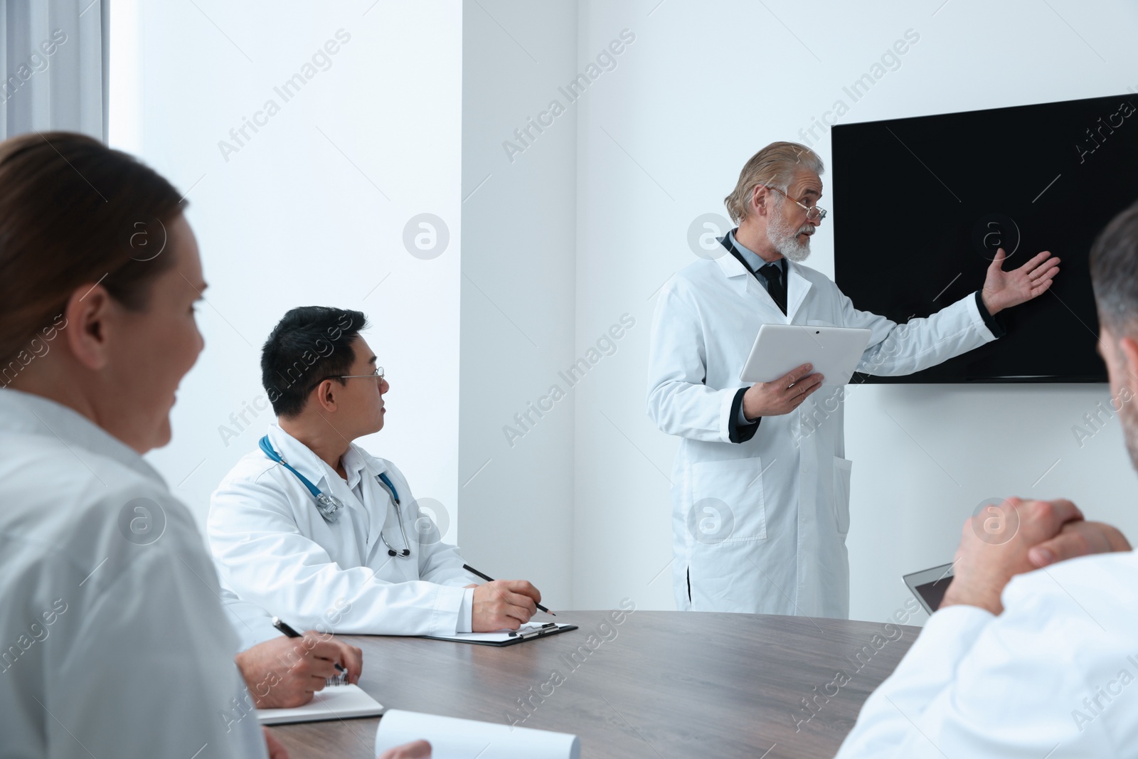 Photo of Team of doctors listening to senior speaker report near tv screen in meeting room. Medical conference