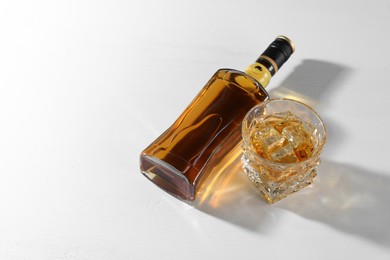 Whiskey with ice cubes in glass and bottle on white table, above view. Space for text