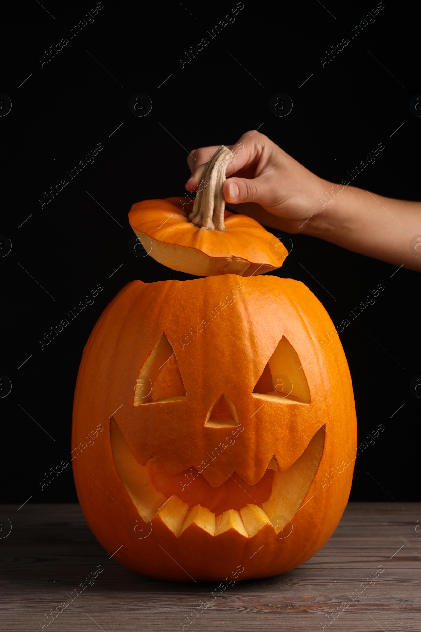 Photo of Woman with scary jack o'lantern made of pumpkin on wooden table against black background, closeup. Halloween traditional decor