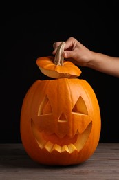 Woman with scary jack o'lantern made of pumpkin on wooden table against black background, closeup. Halloween traditional decor