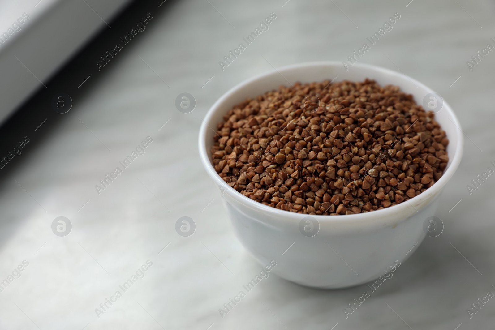 Photo of Bowl with uncooked buckwheat porridge on white marble table, space for text