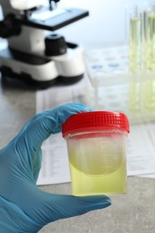 Photo of Doctor holding container with urine sample for analysis at grey table in laboratory, closeup