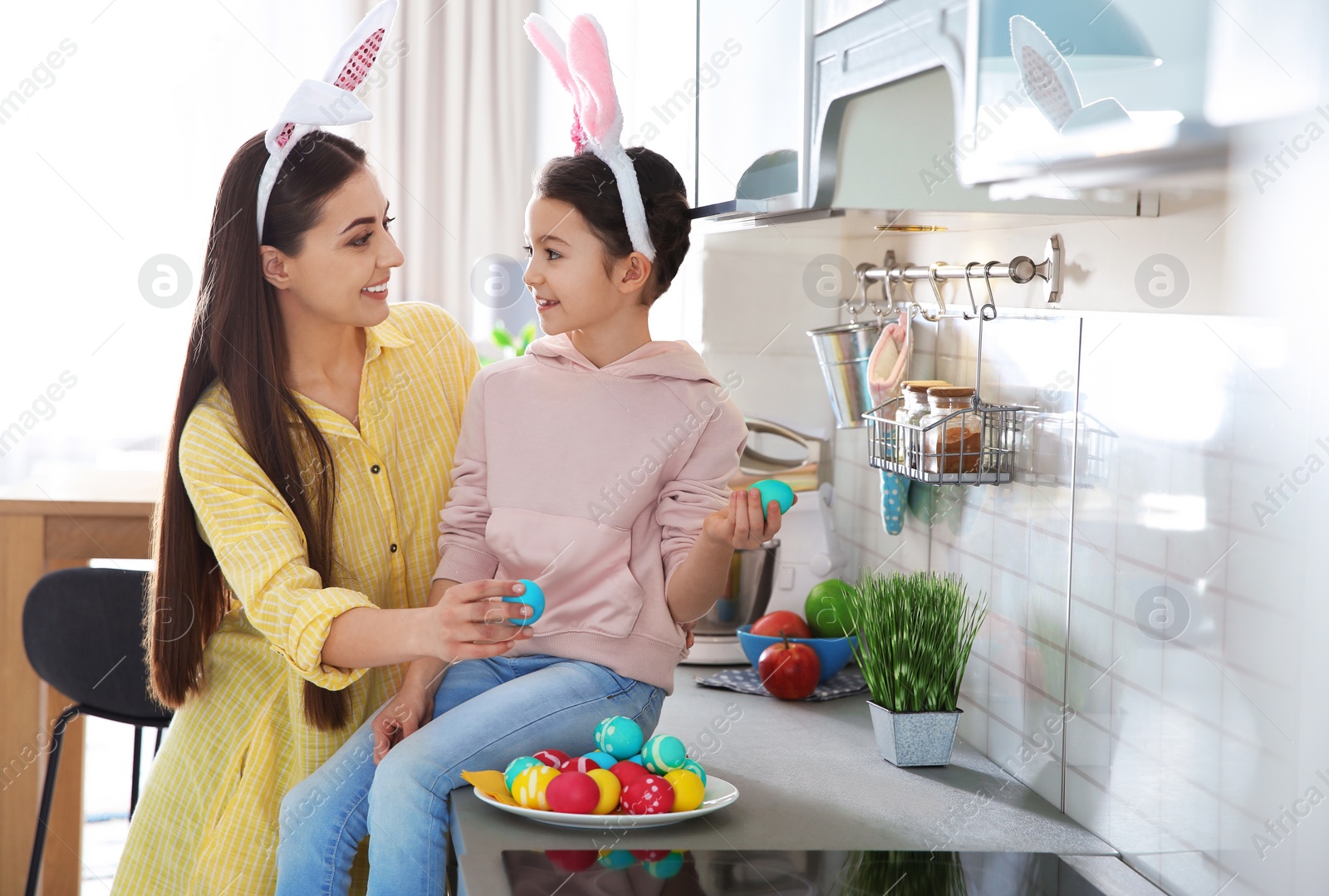Photo of Mother and daughter with bunny ears headbands and painted Easter eggs in kitchen, space for text