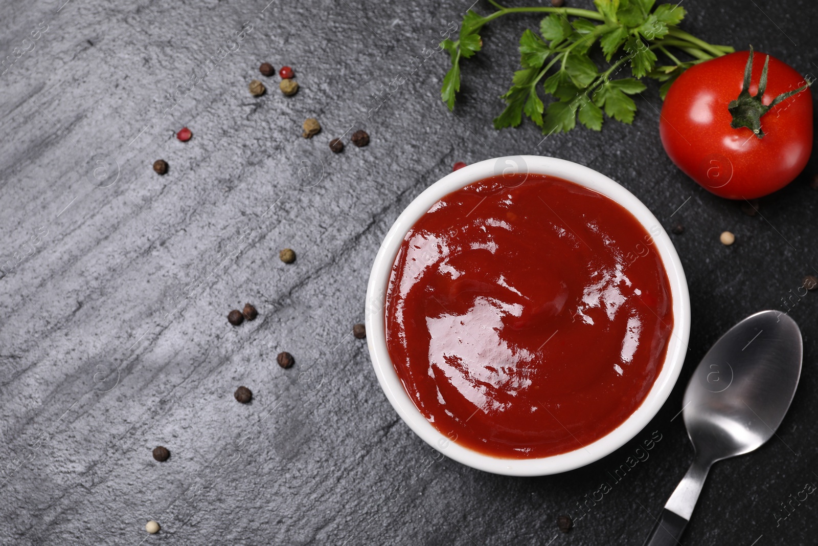 Photo of Organic ketchup in bowl, fresh tomato and spices on black table, flat lay with space for text. Tomato sauce