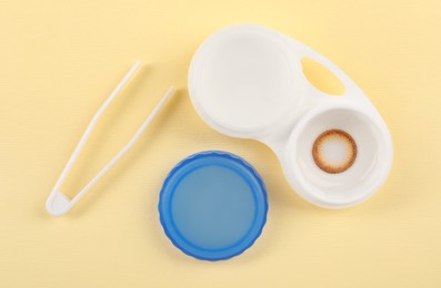 Photo of Case with color contact lenses and tweezers on pale yellow background, flat lay