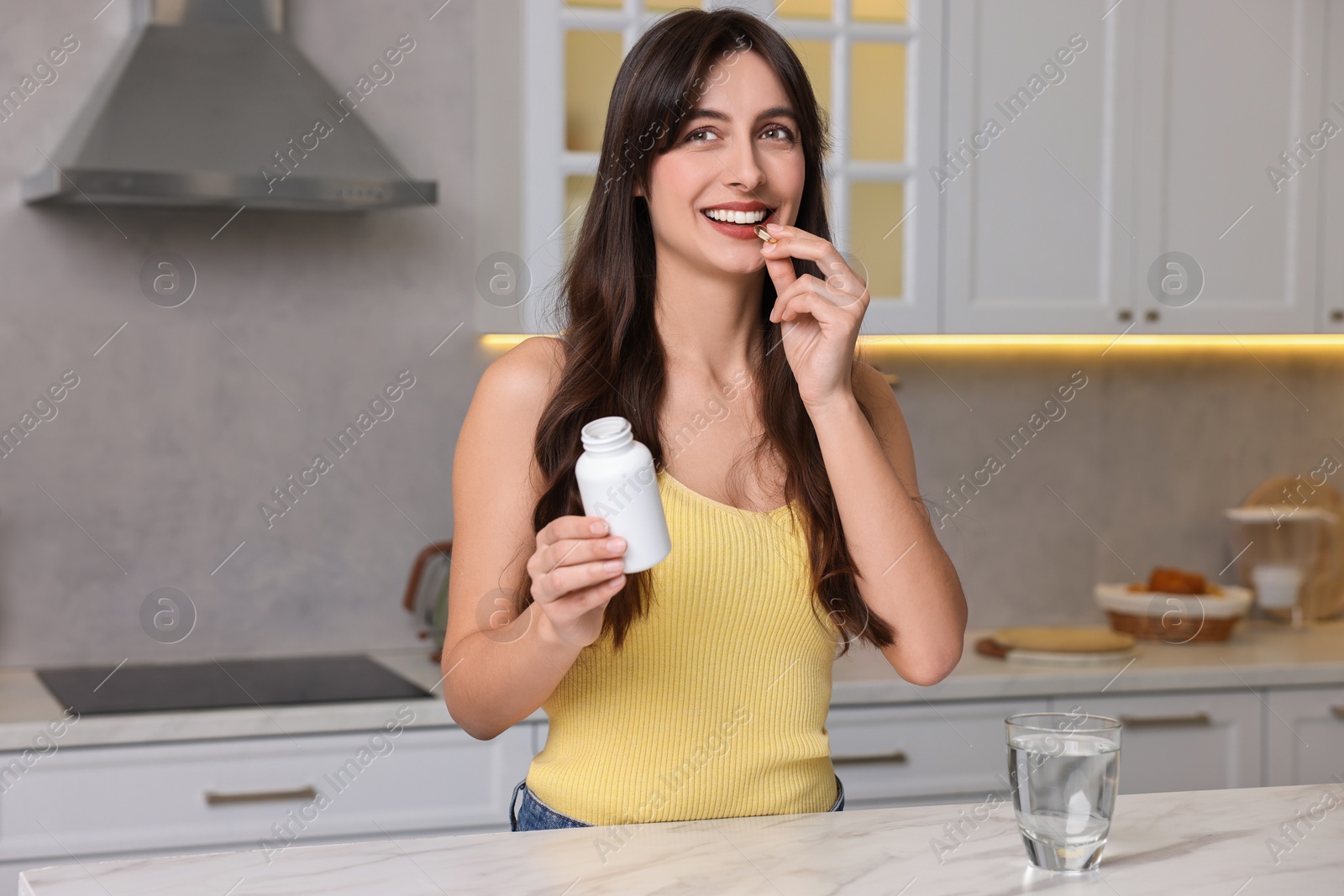 Photo of Beautiful woman taking vitamin pill at white marble table in kitchen