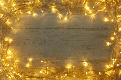 Frame made of glowing Christmas lights on white wooden background, top view. Space for text