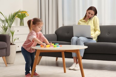 Photo of Woman working remotely at home. Mother talking on smartphone and using laptop while her daughter playing at desk