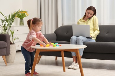 Woman working remotely at home. Mother talking on smartphone and using laptop while her daughter playing at desk