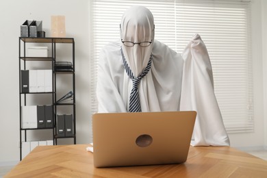 Photo of Overworked ghost. Man covered with white sheet using laptop at wooden table in office