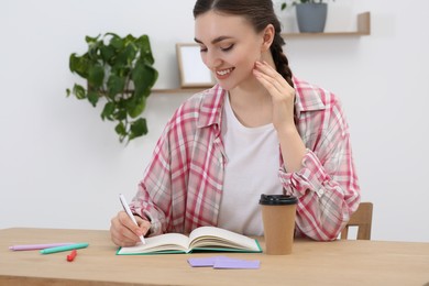 Happy young woman writing in notebook at wooden table indoors