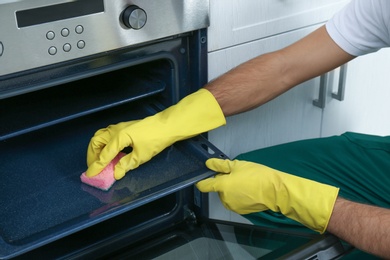 Photo of Male janitor cleaning oven tray with sponge in kitchen, closeup
