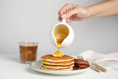 Photo of Woman pouring maple syrup onto delicious pancakes with butter and fried bacon at white wooden table, closeup