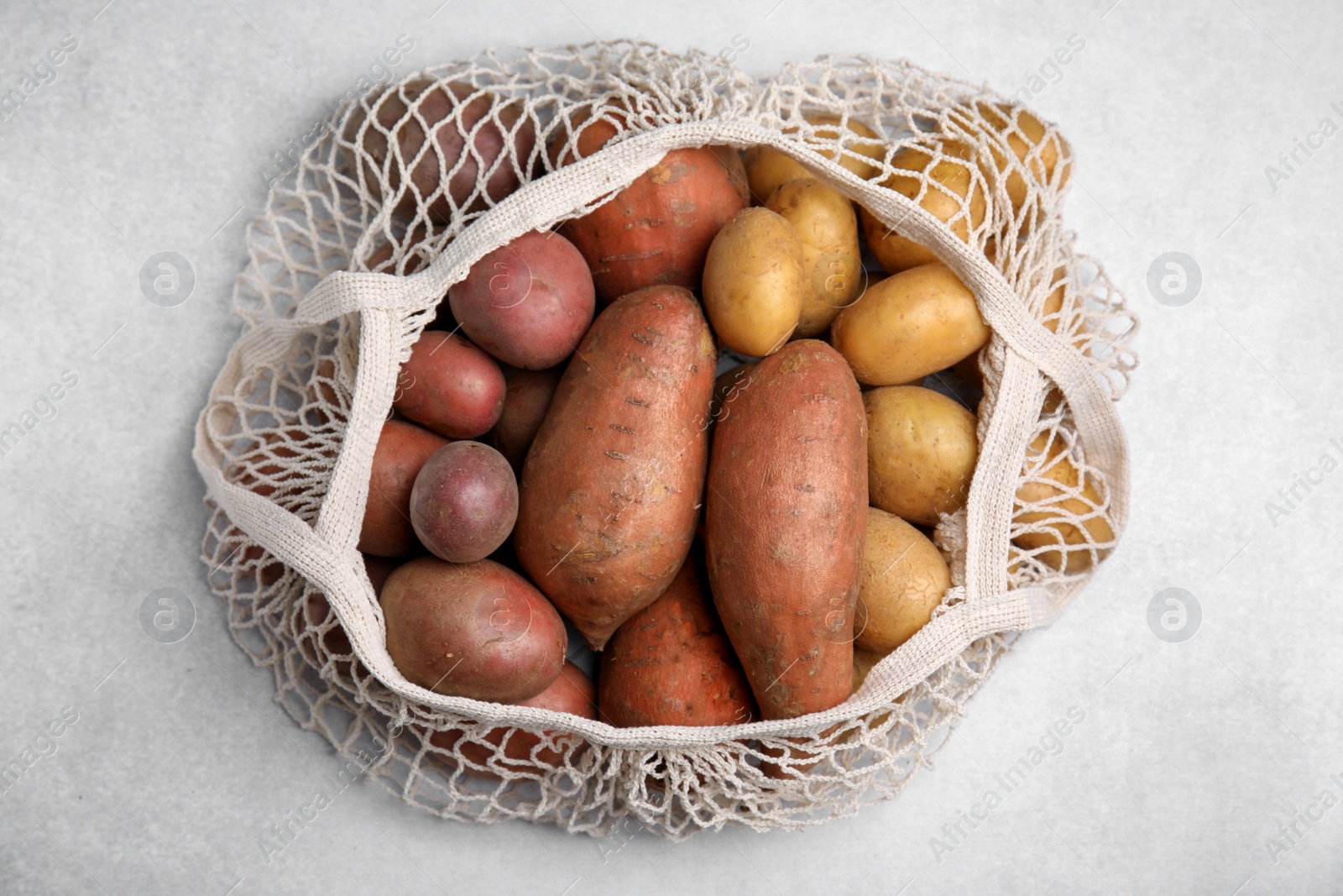 Photo of Different types of fresh potatoes in net bag on light gray table, top view