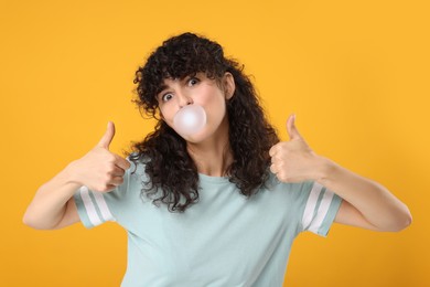 Photo of Beautiful young woman blowing bubble gum and showing thumbs up on orange background