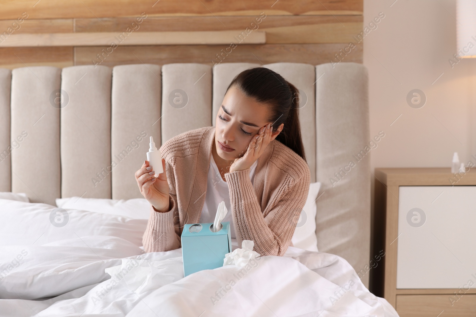 Photo of Sick young woman with napkins and nasal spray in bed at home