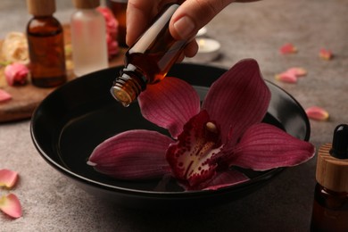 Photo of Woman dripping essential oil into bowl with flower at grey table, closeup. Aromatherapy treatment