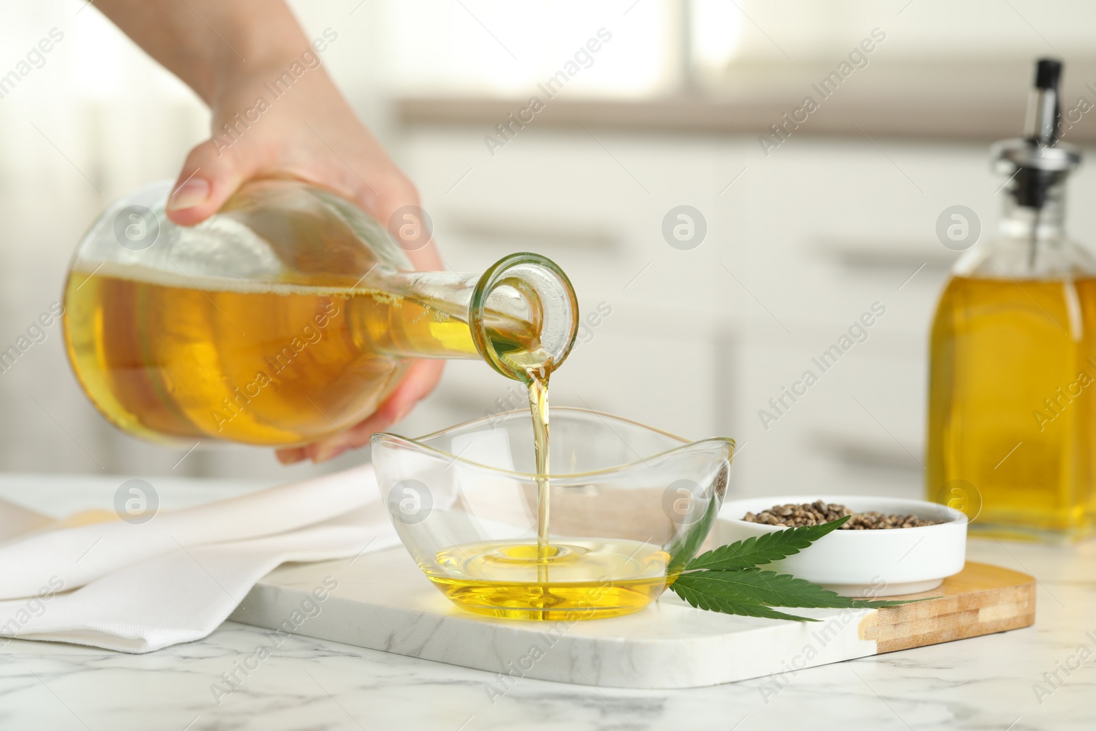 Photo of Woman pouring hemp oil into glass bowl at white marble table, closeup