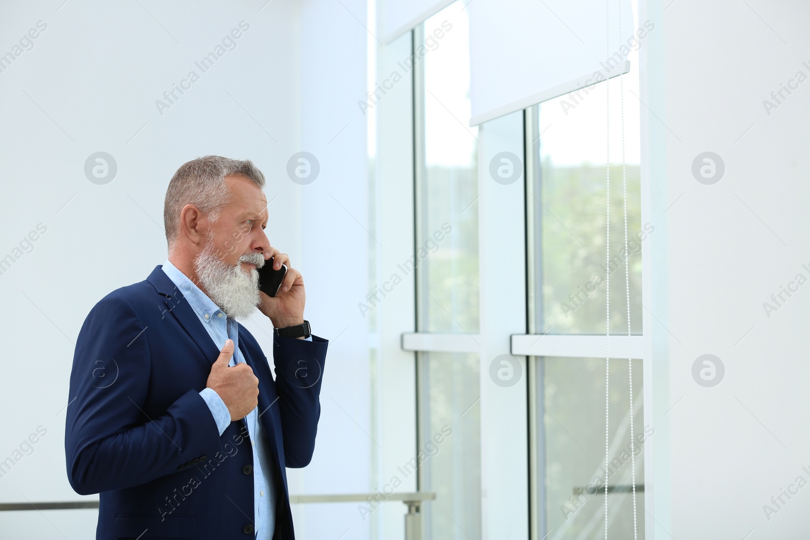 Photo of Portrait of handsome mature man in elegant suit talking on mobile phone near window