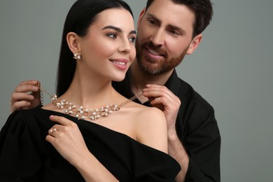 Man putting elegant necklace on beautiful woman against grey background, closeup