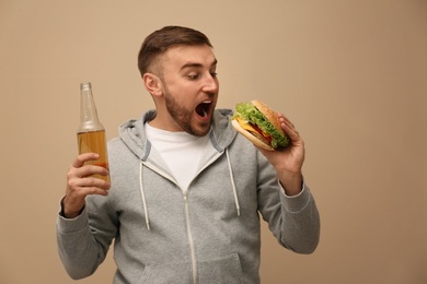 Young man with beer eating tasty burger on color background