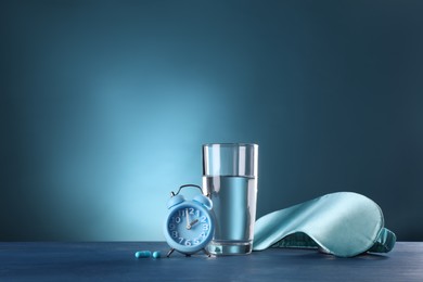 Photo of Alarm clock, soporific pills, sleeping mask and glass of water on blue wooden table, space for text. Insomnia treatment