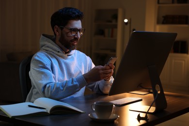 Photo of Home workplace. Happy man using smartphone while working with computer at wooden desk in room at night