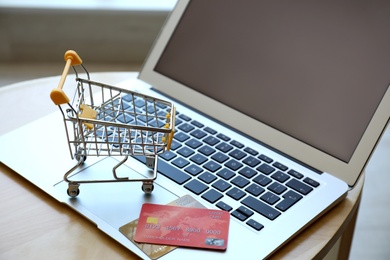 Photo of Internet shopping. Laptop, small cart and credit cards on table indoors, closeup