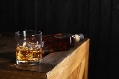 Photo of Whiskey with ice cubes in glass and bottle on wooden crate against black background, space for text