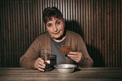 Photo of Poor senior woman with water and bread at table against wooden wall