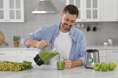 Photo of Happy man pouring delicious smoothie into glass at white marble table in kitchen