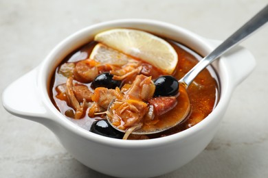 Photo of Meat solyanka soup with sausages, olives, vegetables and spoon in bowl on white textured table, closeup