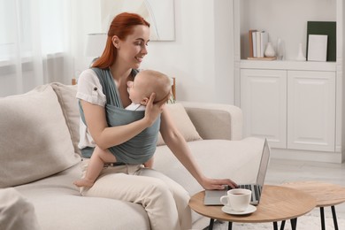 Photo of Mother holding her child in sling (baby carrier) while using laptop at home