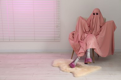 Photo of Glamorous ghost. Woman in pink sheet and high heel shoes on armchair indoors, space for text